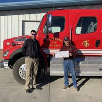 two men standing in from of a red truck. One is holding a large check