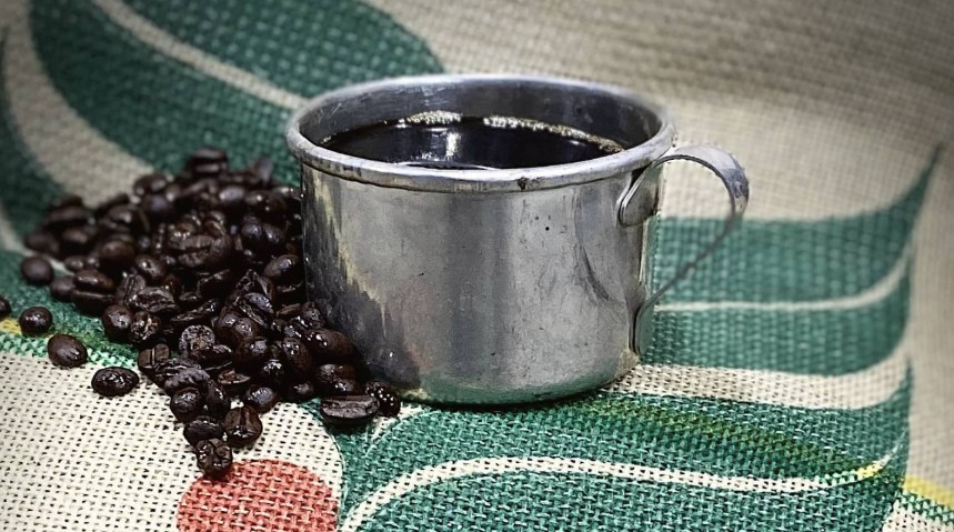 Bates Bros Coffee – Father’s Day Gift Ideas Freshly brewed coffee in a metal camp mug surrounded by roasted Bates Bros Coffee beans on a burlap coffee sack.
