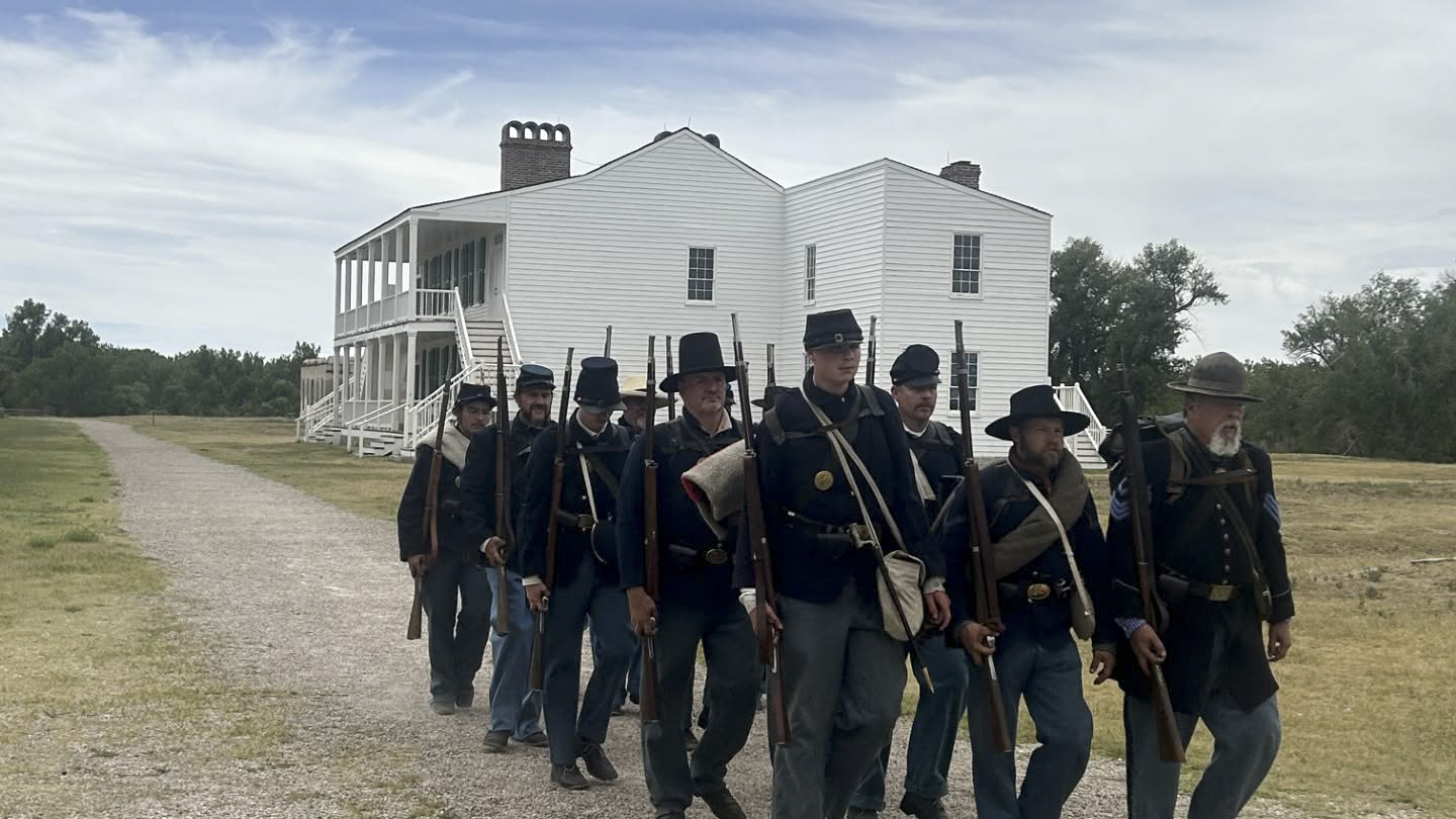 Living history reenactors portraying U.S. Army soldiers march past the historic Old Bedlam building during Living History Weekend at Fort Laramie National Historic Site.
